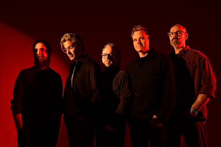 Indie rock band Death Cab for Cutie standing together in dark outfits bathed in dramatic red lighting.